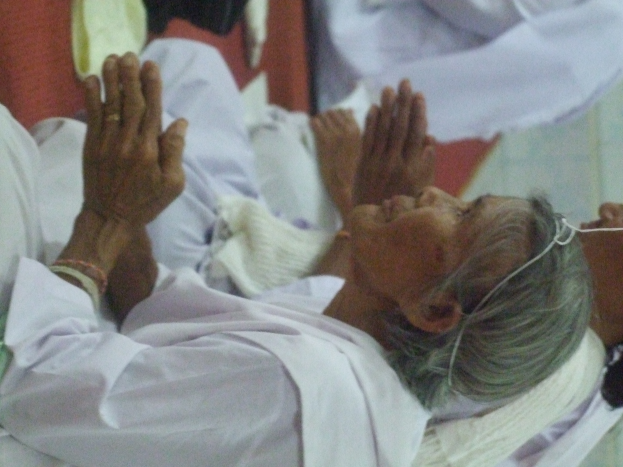 Eine ältere Frau liegt in einem Krankenhausbett, die Hände gefaltet, und sieht besorgt aus, die Augen geschlossen und den Kopf auf den Händen ruhend, umgeben von ein paar Menschen im Hintergrund.