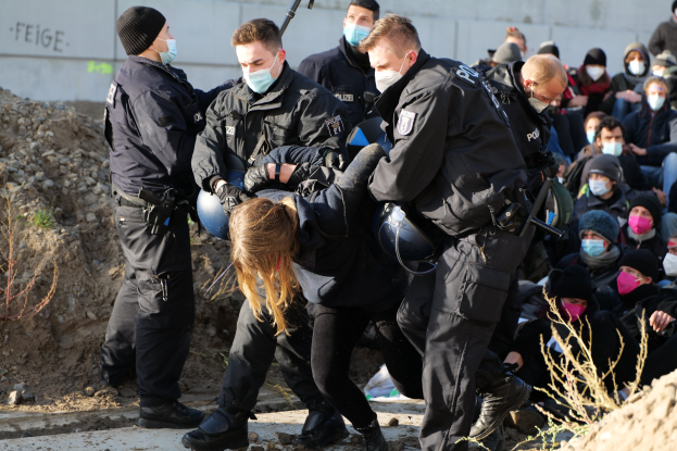 Maskierte Gruppe in Mützen und mit Waffen umringt eine Frau, die von der Polizei eskortiert wird.