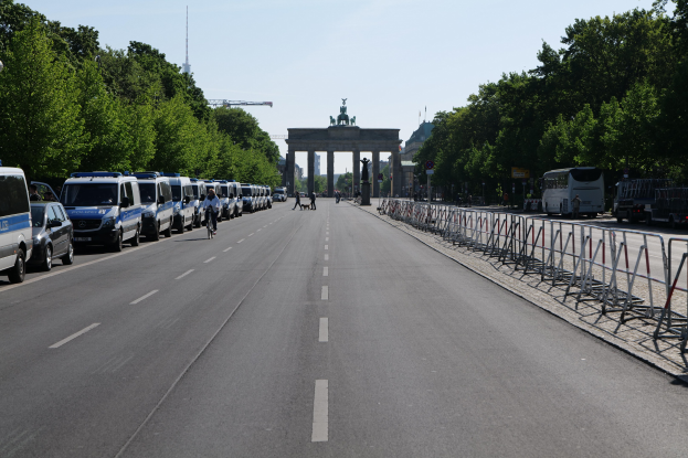 Lange Reihe von Polizeiwagen entlang einer Straße vor dem Brandenburger Tor geparkt, mit Radfahrern und Fußgängern, Absperrungen, Bäumen und einem Bogen mit Statuen im Hintergrund.