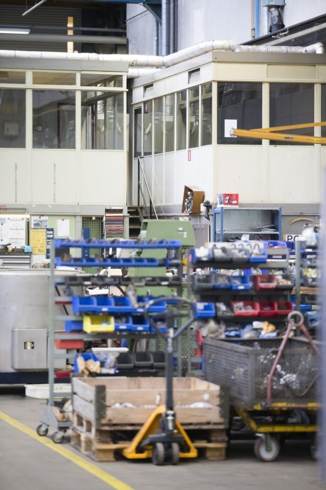 Lagerinneres mit verschiedenen Geräten einschließlich fahrbaren Wagen, Schränken und einer Box, mit einer Glasfensterwand und Rohren im Hintergrund.