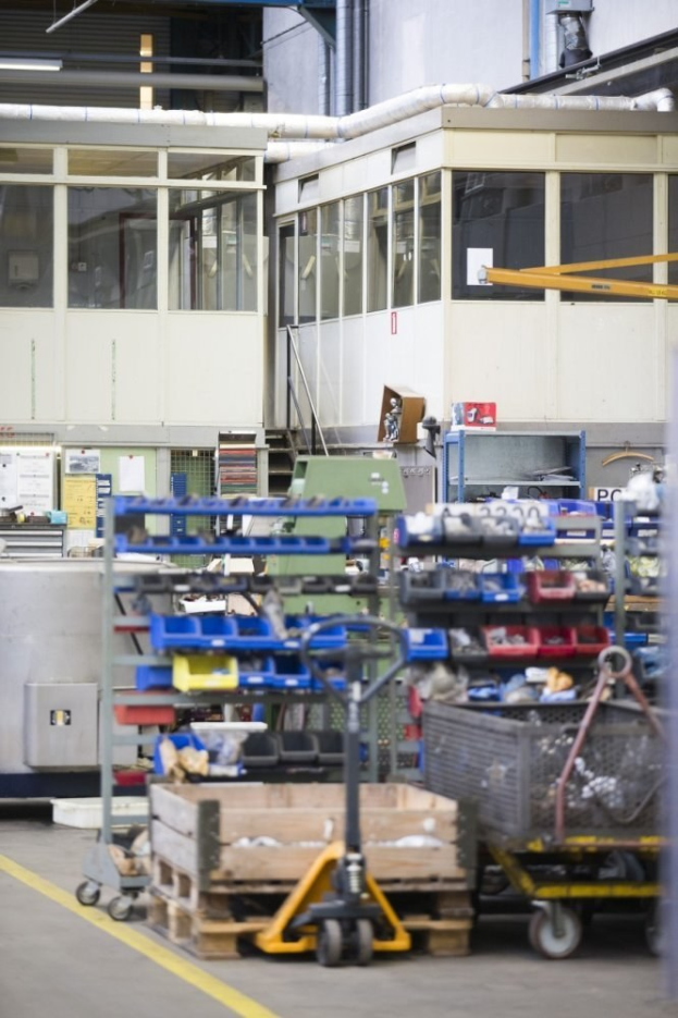 Lagerinneres mit verschiedenen Geräten einschließlich fahrbaren Wagen, Schränken und einer Box, mit einer Glasfensterwand und Rohren im Hintergrund.