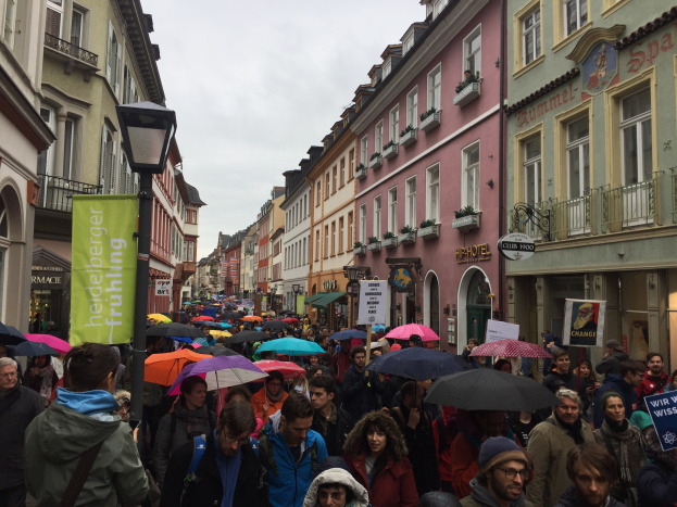 Große Gruppe von Menschen, die während einer Klimademonstration in Deutschland die Straße entlanggehen, viele mit Schirmen, einige mit Taschen und Schildern, sowie Gebäude und einen Laternenpfahl im Hintergrund.