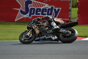 Eine Person in Helm und Schutzkleidung fährt ein Motorrad auf einer Rennstrecke, umgeben von Gras und einer Tafel mit Text im Hintergrund.