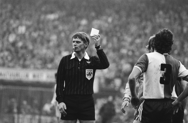 Schwarzes Bild eines Schiedsrichters mit roter Karte vor einer Menge in Fußballuniformen und einem Zaun im Hintergrund.