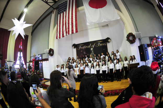 Gruppe von Menschen auf einer Bühne mit Weihnachtsdekoration, einem Podium, Lautsprechern, einer Texttafel, Vorhängen, Fahnen und Deckenleuchten vor einem Publikum mit Kameras und Handys.