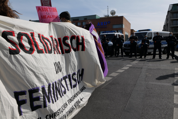 Gruppe von Menschen marschiert auf einer Straße und hält ein 'Solidarität und Feminismus'-Schild, mit parkenden Fahrzeugen, Gebäuden, einer Schüsselantenne und einem klaren blauen Himmel im Hintergrund.