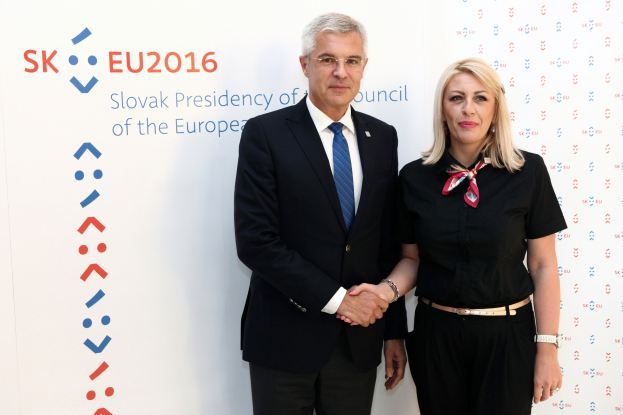 Zwei Personen, die nebeneinanderstehen und lächeln, mit einer Tafel im Hintergrund, auf der "sk eu 2016" steht; der Mann trägt einen schwarzen Blazer, ein weißes Hemd, eine blaue Krawatte und schwarze Hosen, und die Frau trägt eine Brille.