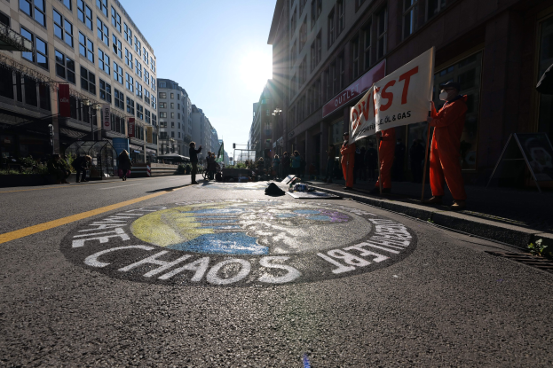 Eine Gruppe von Menschen hält Protestschilder und Banner auf einer Stadtstraße mit Gebäuden, Pflanzen und einer "Chaos"-Straßenmalerei im Hintergrund.