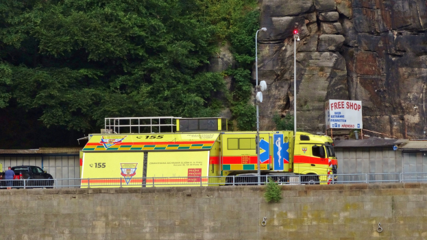 Ambulanz auf der Seite einer Straße neben einer Klippe geparkt, mit verschiedenen Straßenobjekten im Vordergrund und Bäumen und einem Auto im Hintergrund.