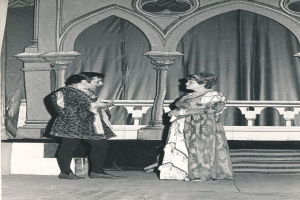 Ein Schwarz-Weiß-Foto eines Mannes und einer Frau, die auf einer Bühne vor einer Wand mit Säulen und einem Vorhang stehen, beide in Abendgarderobe gekleidet.