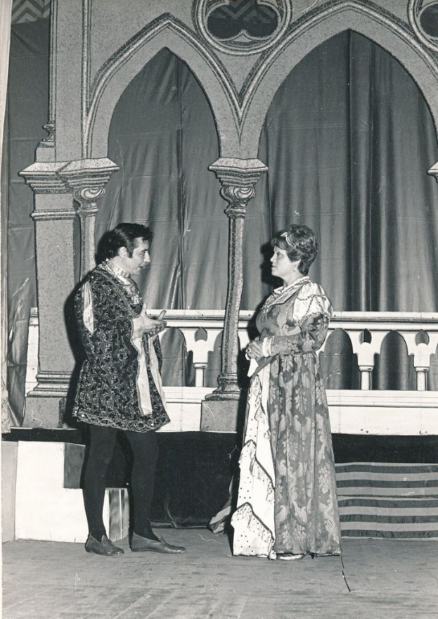 Ein Schwarz-Weiß-Foto eines Mannes und einer Frau, die auf einer Bühne vor einer Wand mit Säulen und einem Vorhang stehen, beide in Abendgarderobe gekleidet.