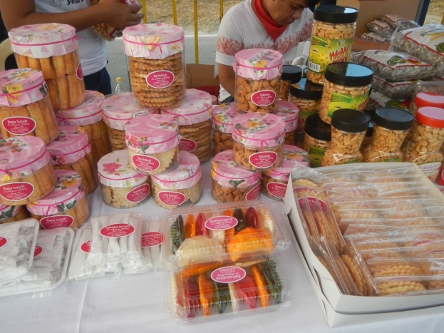 Eine Gruppe von Menschen steht um einen Tisch mit verschiedenen Lebensmitteln auf einem Food-Festival, mit einem Geländer und einer Straße im Hintergrund.