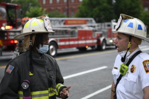 Zwei Feuerwehrleute in Helmen, einer mit Maske, unterhalten sich auf einer Straße mit Fahrzeugen, Bäumen, Gebäuden und einem Mast im Hintergrund.