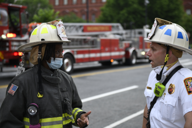 Zwei Feuerwehrleute in Helmen, einer mit Maske, unterhalten sich auf einer Straße mit Fahrzeugen, Bäumen, Gebäuden und einem Mast im Hintergrund.