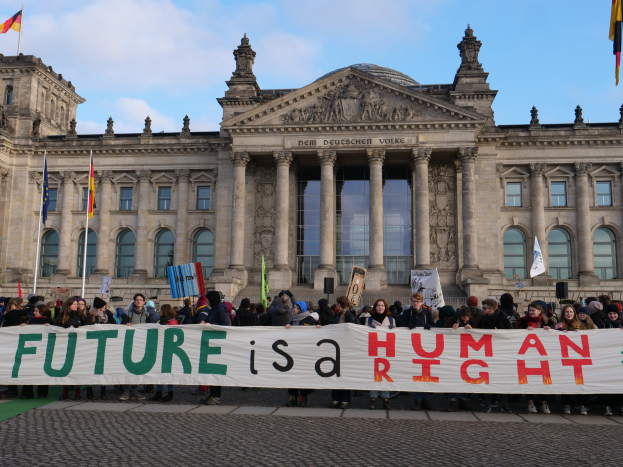 Eine Gruppe von Menschen hält ein Transparent mit der Aufschrift "Zukunft ist ein Menschenrecht" vor dem Reichstaggebäude in Berlin, Deutschland, das mit Fenstern, Säulen und Bögen verziert ist und von Fahnenstangen mit Fahnen umgeben ist, unter einem bewölkten Himmel.