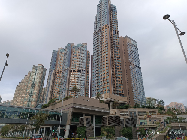 Eine Stadtstraße mit hohen Gebäuden, Fahrzeugen, Laternenmästen, einer Brücke, Bäumen, Pflanzen und einem bewölkten Himmel, mit einem Wohnhaus im Vordergrund.