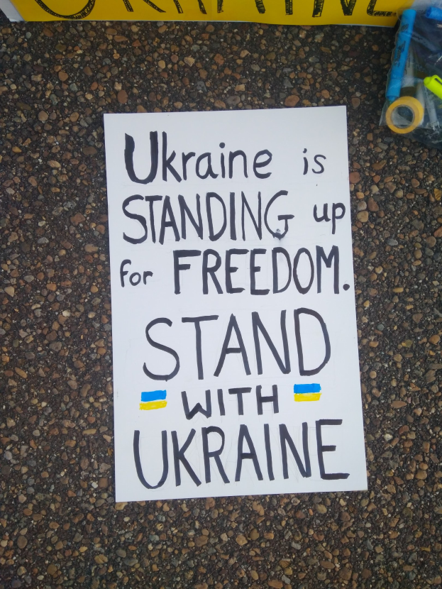 Weißer Zettel mit "Ukraine steht für Freiheit, steht mit Ukraine"-Text auf dem Boden vor einem gelben Banner, mit einer Plastikhülle mit verschiedenen Gegenständen rechts.