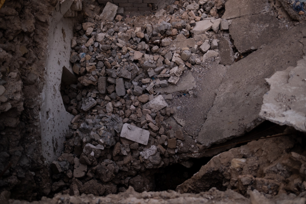 Zerstörtes Gebäude mit einem großen Loch im Boden, umgeben von Schutt und einer sichtbaren Rohrleitung auf der linken Seite, was auf Erdbebenschäden hinweist.