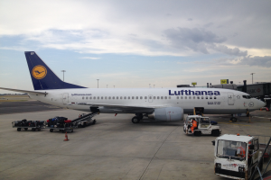 Ein Lufthansa Airbus A320-200 steht auf dem Frankfurter Flughafen, umgeben von verschiedenen Bodenfahrzeugen, Wagen, Absperrgittern, Pfählen und Texttafeln unter einem bewölkten Himmel.