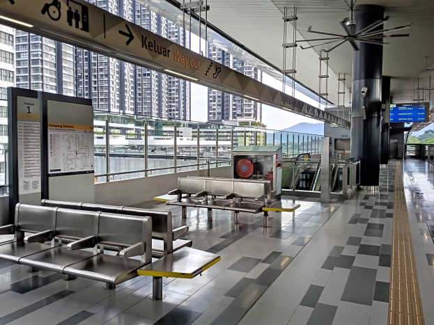 Leerer Flughafen-Wartebereich mit mehreren Bänken, Informationsanzeigen, Säulen, einem Deckenventilator und Leuchten, vor einem Hintergrund aus Gebäuden, Bäumen, Wasser und einem klaren blauen Himmel.