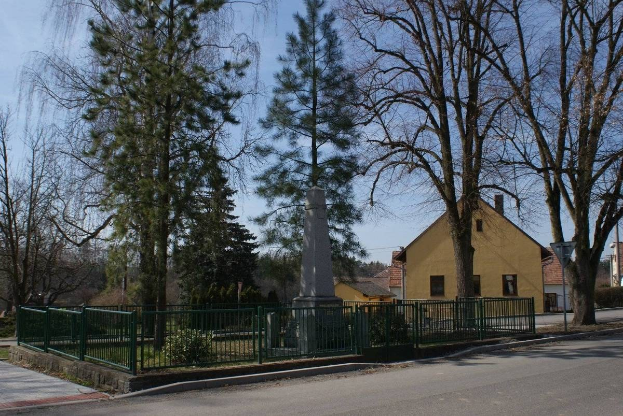 Ein Holocaust-Denkmal steht in der Mitte einer Straße, umgeben von einem Metallzaun, Grünflächen, Häusern, einem Straßenmast und einem bewölkten Himmel.