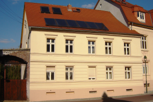 Gebäude mit Solarpaneelen auf dem Dach, umgeben von Straßeninfrastruktur, Bäumen und bewölktem Himmel.