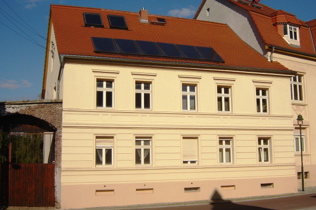 Gebäude mit Solarpaneelen auf dem Dach, umgeben von Straßeninfrastruktur, Bäumen und bewölktem Himmel.