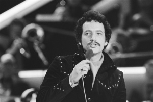 Ein als Elvis Presley identifizierter Mann hält ein Mikrofon während einer Live-Performance in einem Schwarz-Weiß-Foto mit unscharfem Hintergrund.