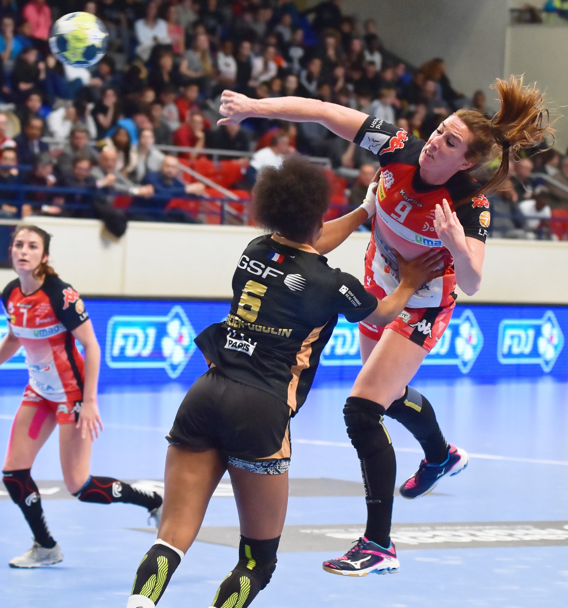 Eine Gruppe von Frauen, die Handball auf einem Feld mit einem Ball in der Luft spielen, in einer Arena mit Zuschauern im Hintergrund.