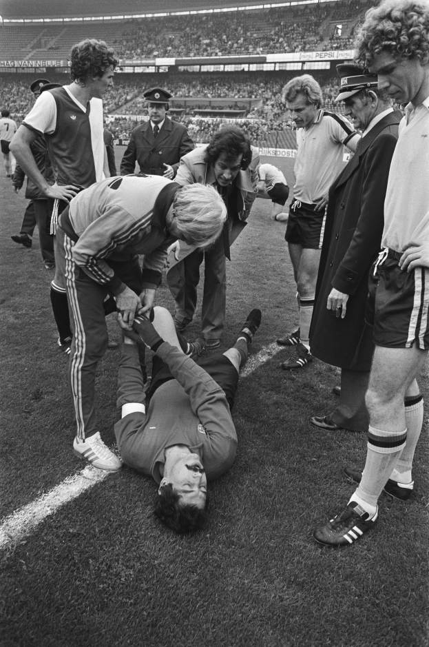 Schwarzes und weißes Bild eines Fußballspielers, der auf dem Boden liegt und von einer Gruppe Männer umgeben ist, mit einer Stadionkulisse im Hintergrund; der Spieler scheint verletzt zu sein.