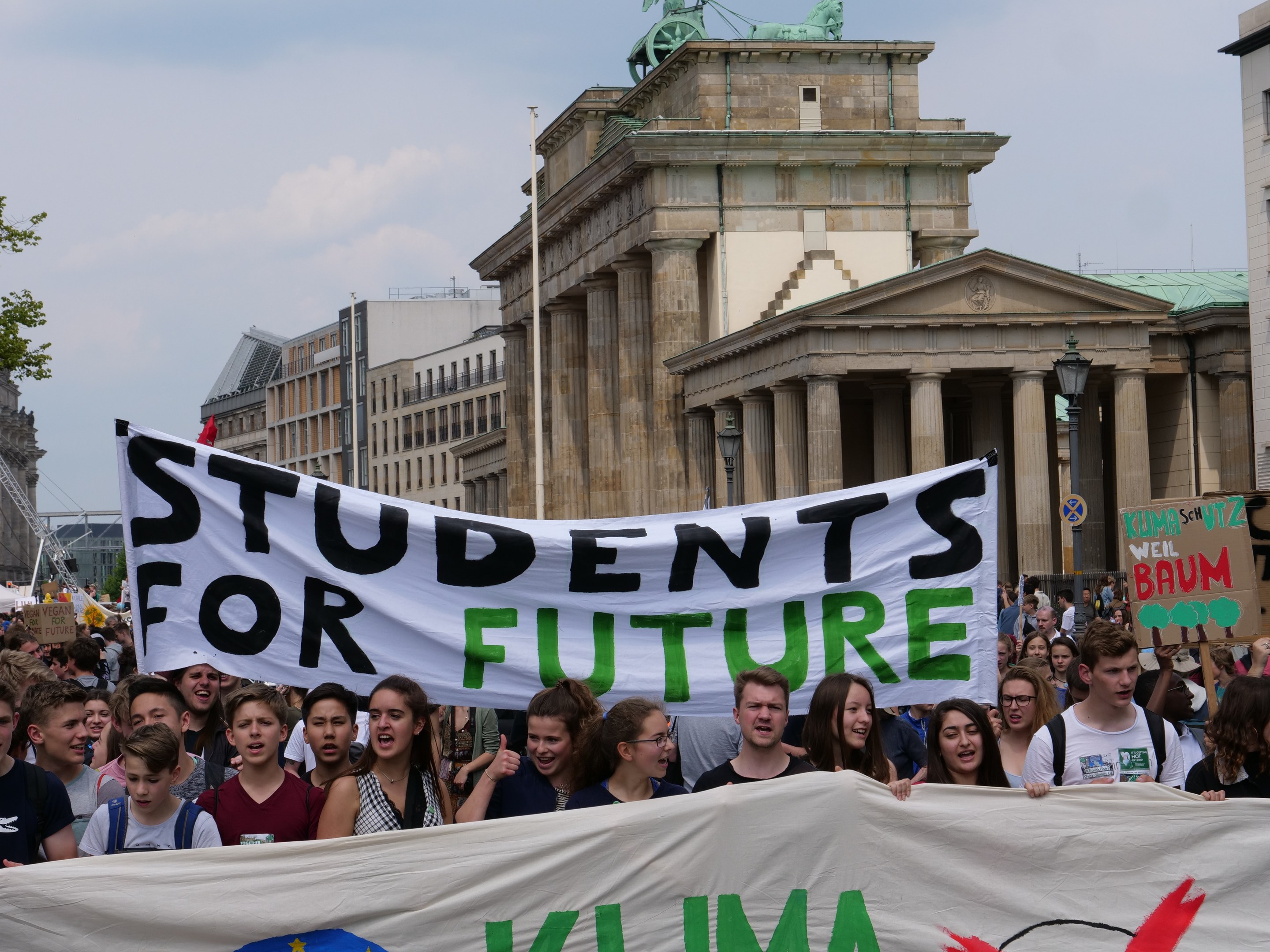 Gruppe von Schülern marschiert in Berlin mit einem leuchtend bunten "Students for Future"-Schild vor einem Hintergrund von Gebäuden, Bäumen und Himmel.