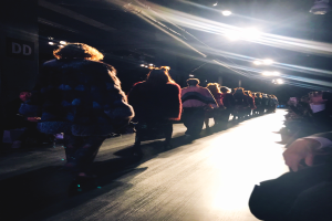 Eine Gruppe von Menschen läuft über den Laufsteg einer Fashion Show, beleuchtet von hellen Scheinwerfern, während Zuschauer auf beiden Seiten die Show beobachten.