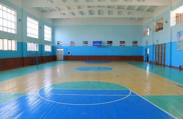 Eine große Turnhalle mit einem blauen und weißen Boden, die einen Basketballplatz, ein Netz mit Pfosten, eine Tür, Fenster, Plakate an den Wänden, ein Geländer und ein Dach zeigt.