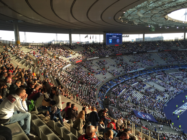 Eine große Menschenmenge in einem Stadion schaut ein Fötballspiel zu, mit einer Bühne auf der rechten Seite, Fahnen, Masten, einem Bildschirm und der Allianz Arena in München im Hintergrund.
