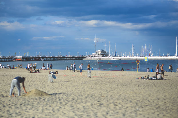 Eine Gruppe von Menschen, die Volleyball auf einem sandigen Strand mit einem Netz, Booten und einer Brücke mit Geländern im Hintergrund unter einem bewölkten Himmel spielen.