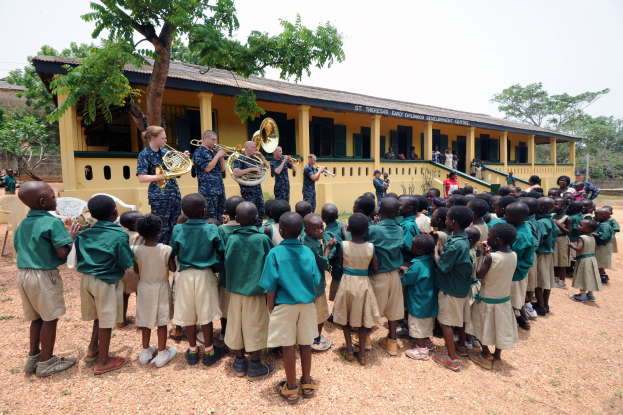 Eine Gruppe von Kindern steht vor einem Gebäude mit Fenstern, Säulen und einer Treppe, einige halten Musikinstrumente, mit Bäumen und einer Wand im Hintergrund unter einem bewölkten Himmel am St. Theresa Early Childhood Development Centre in Uganda.