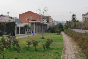 Eine Stadtstraße mit einem zentralen Fußweg, gesäumt von Bänken, umgeben von Gebäuden, Straßenlaternen, Bäumen und Fahrzeugen unter einem klaren blauen Himmel.