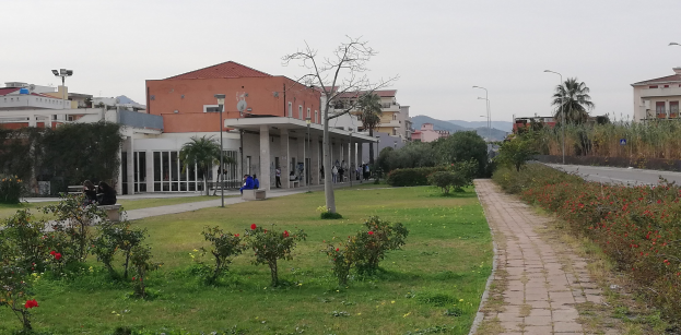 Eine Stadtstraße mit einem zentralen Fußweg, gesäumt von Bänken, umgeben von Gebäuden, Straßenlaternen, Bäumen und Fahrzeugen unter einem klaren blauen Himmel.