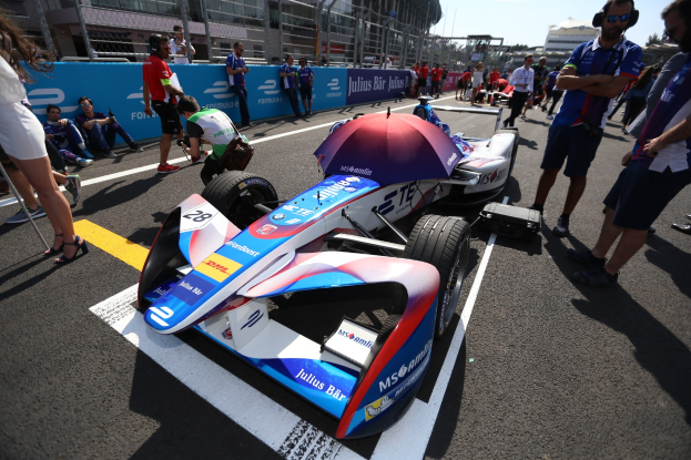Formula E-Rennwagen mit weißer, blauer, roter und schwarzer Lackierung, der auf einer Strecke neben Menschen geparkt ist, mit einem Schirm im Vordergrund und Renninfrastruktur im Hintergrund unter einem klaren Himmel.