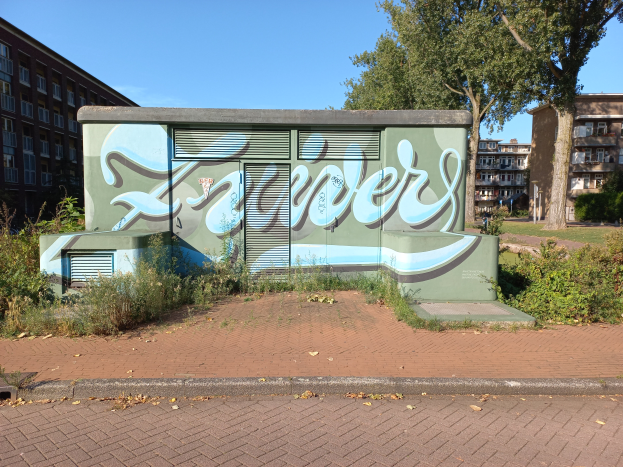 Ein grünes Gebäude mit Graffiti an seiner Seite, umgeben von Pflanzen und Bäumen, mit anderen Gebäuden im Hintergrund und einem klaren blauen Himmel darüber.