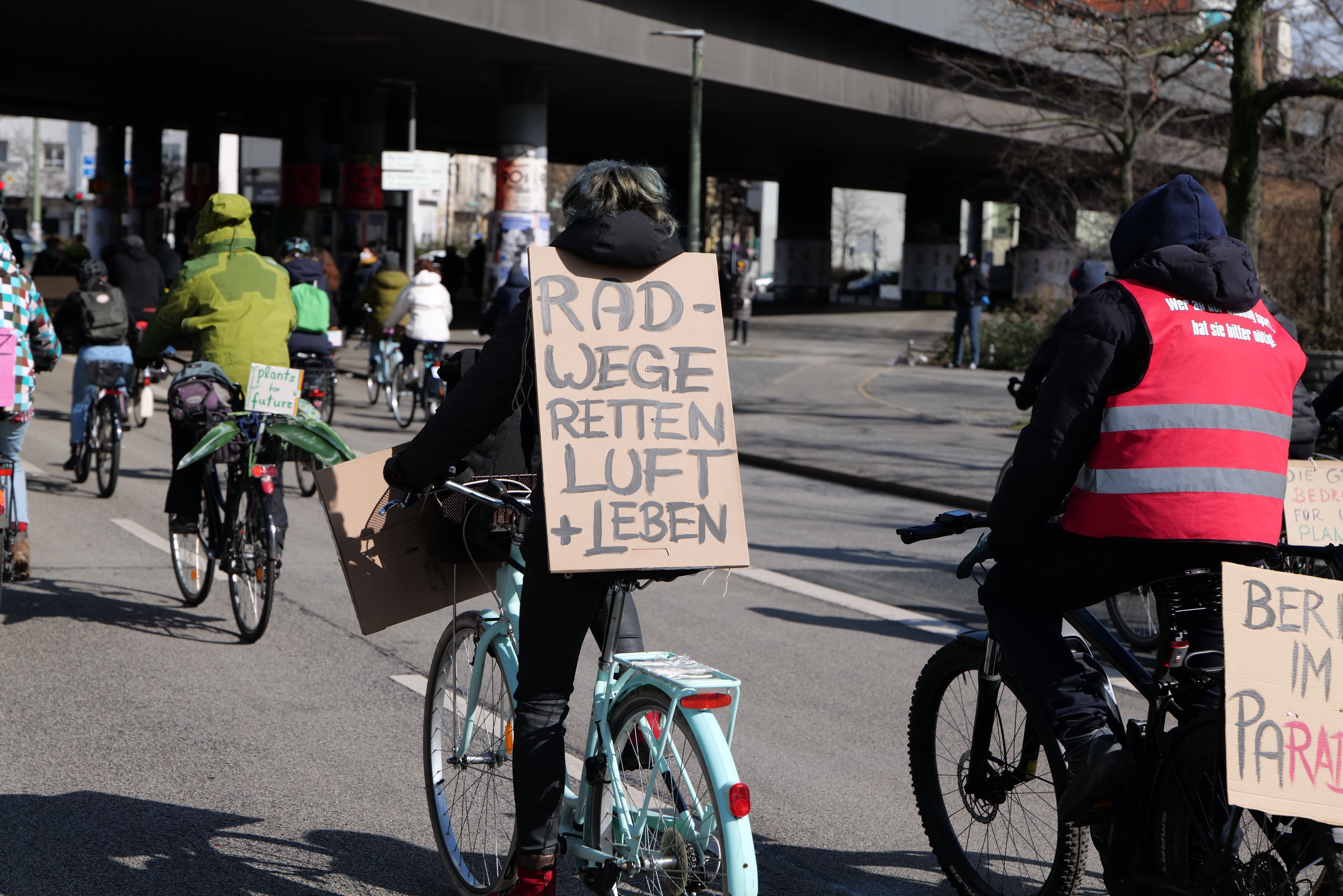 Gruppe von Menschen, die Fahrräder auf einer Straße mit einer Brücke im Hintergrund fahren, Bäume säumen die Straße und Gebäude sind in der Ferne zu sehen, sie halten Schilder mit verschiedenen Botschaften.