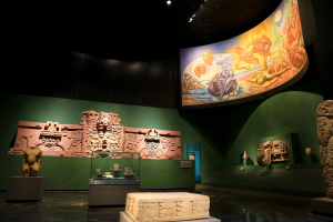Ein Museumsraum mit verschiedenen Artefakten, darunter Skulpturen, Tischgegenstände, eine Texttafel, eine Wandmalerei und Deckenbeleuchtung im Mexiko-Stadt-Museum der Anthropologie.