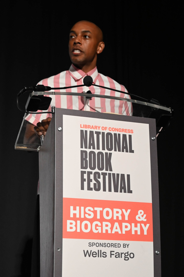 Mann, der an einem Rednerpult mit einem Mikrofon während einer Abendveranstaltung spricht, mit einem Schild im Hintergrund, auf dem "National Book Festival History & Biography" steht.
