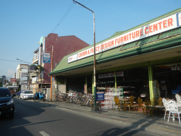 Eine belebte Stadtstraße mit fahrenden Fahrzeugen, parkenden Fahrrädern, Gebäuden mit Schildern, Laternen und Stromleitungen am Himmel, mit einem Möbelgeschäft im Vordergrund.