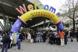 Eine Gruppe von Menschen steht vor einem Willkommensbogen auf einem Outdoor-Event mit Zelten, Bäumen und Gebäuden unter einem klaren blauen Himmel, einige tragen Taschen.