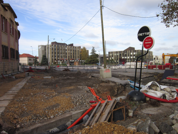 Baustelle mit verstreutem Material, Equipment, Hinweisschildern, Absperrungen, Fahrzeugen, Bäumen, Gebäuden, Laternenmasten und einem bewölkten Himmel im Hintergrund.