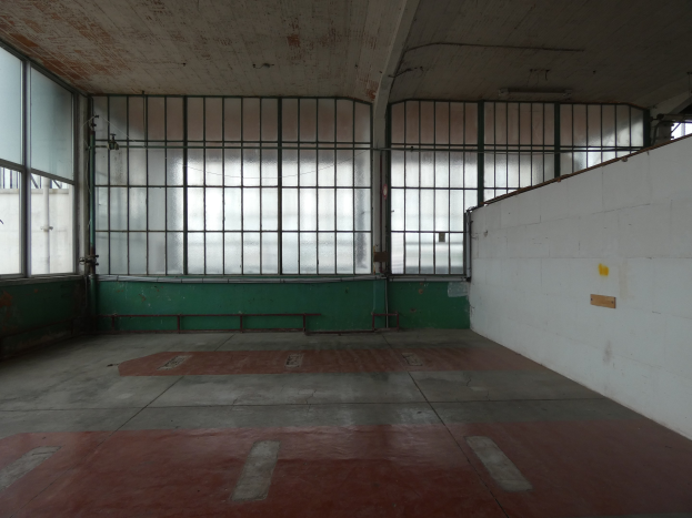 Leeres Gefängniszelle mit rotem Boden, weißer Wand auf der rechten Seite, Metallstangen und Fenstern im Hintergrund.