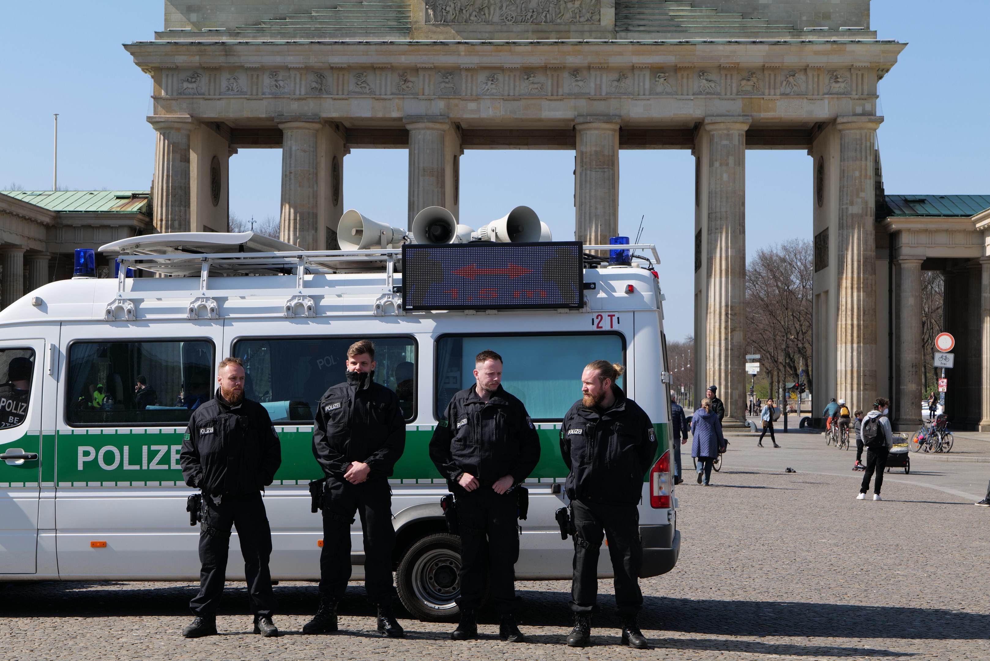 Eine Gruppe von Polizisten in schwarzen Uniformen vor dem Brandenburger Tor in Berlin, mit einem weißen und grünen Fahrzeug im Vordergrund und Fußgängern und Radfahrern im Hintergrund.