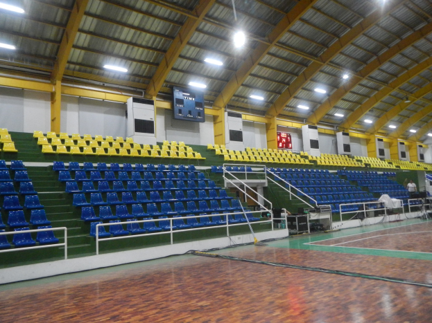 Großer Hallensportplatz mit Holzboden, blauen und gelben Sitzplätzen, Geländern, Treppen mit Geländern, Wandbildschirmen, Deckenleuchten und einigen Menschen.