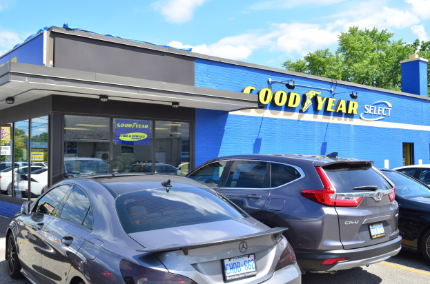 Außensicht einer Goodyear Select-Autohändlung mit parkenden Fahrzeugen, Beschriftung auf dem Gebäude, Bäumen im Hintergrund und einer bewölkten Himmel.